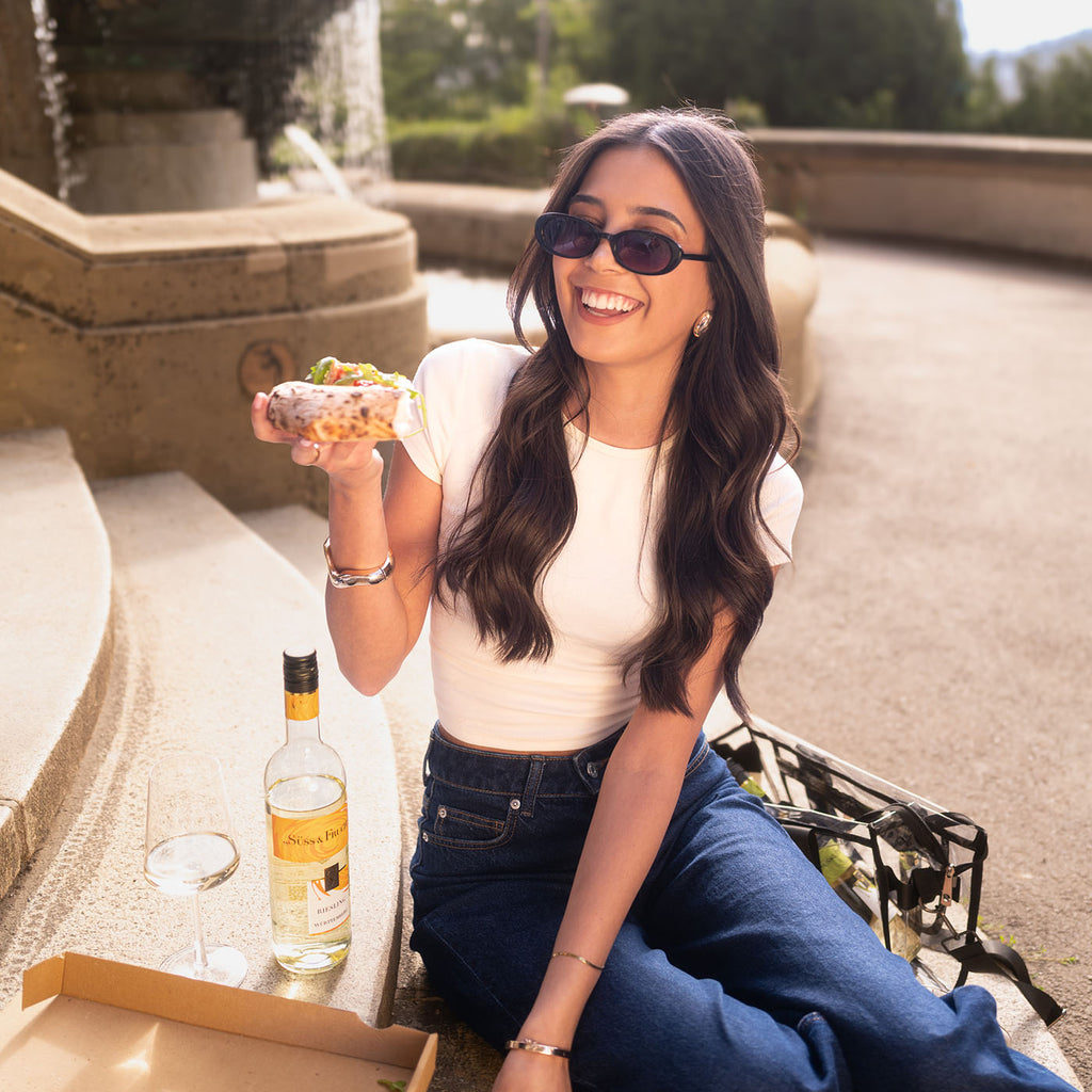 Lachende Frau isst am Brunnen einen Wrap neben einer Flasche Süss & Fruchtig Riesling