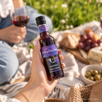 Hand hält eine Flasche Süss & Fruchtig Samtrot von WZG bei einem Picknick mit Rotweinglas und Snacks im Grünen.