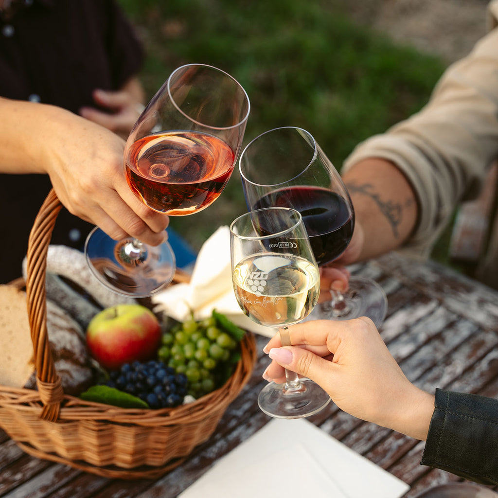 Mehrere Personen stoßen bei einem Picknick mit Wein aus dem Sortiment der WZG über einem Korb mit Snacks an.