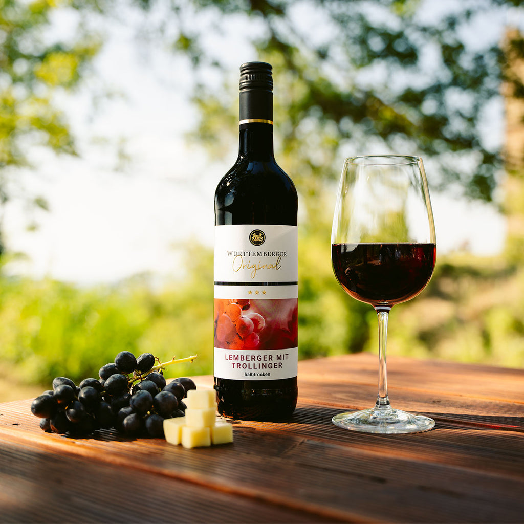 Flasche Lemberger mit Trollinger Rotwein der WZG mit gefülltem Rotweinglas auf einem Holztisch im Grünen.
