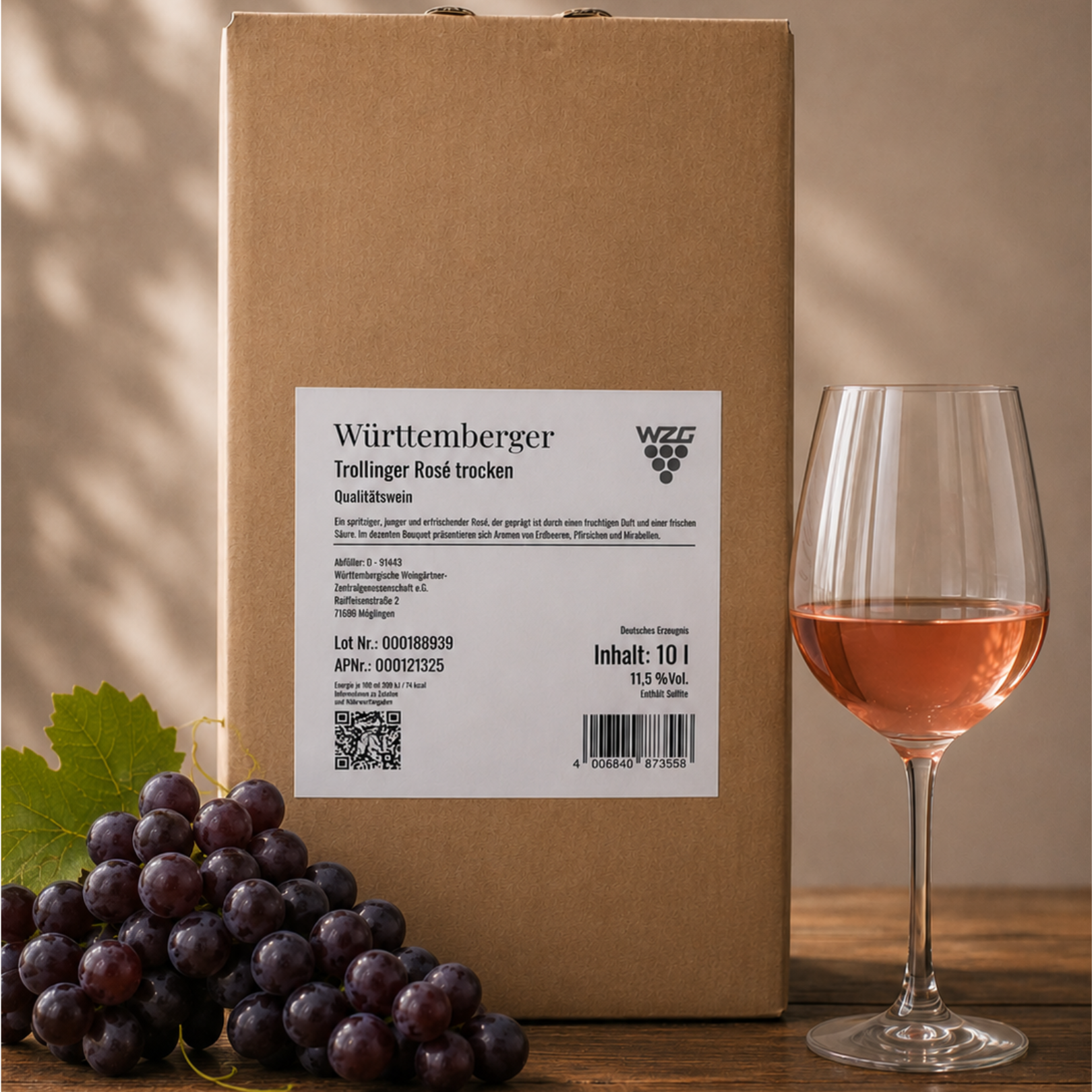 Württemberger Trollinger Rosé trocken im 10 Liter Bag in Box mit gefülltem Roséweinglas und dunklen Trauben auf einem Holztisch.